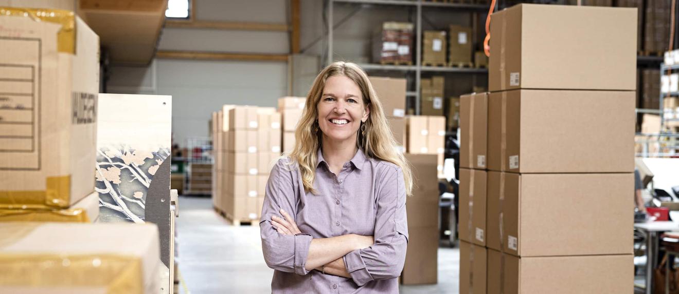 A woman with long blonde hair stands smiling with her arms crossed in a warehouse full of boxes.