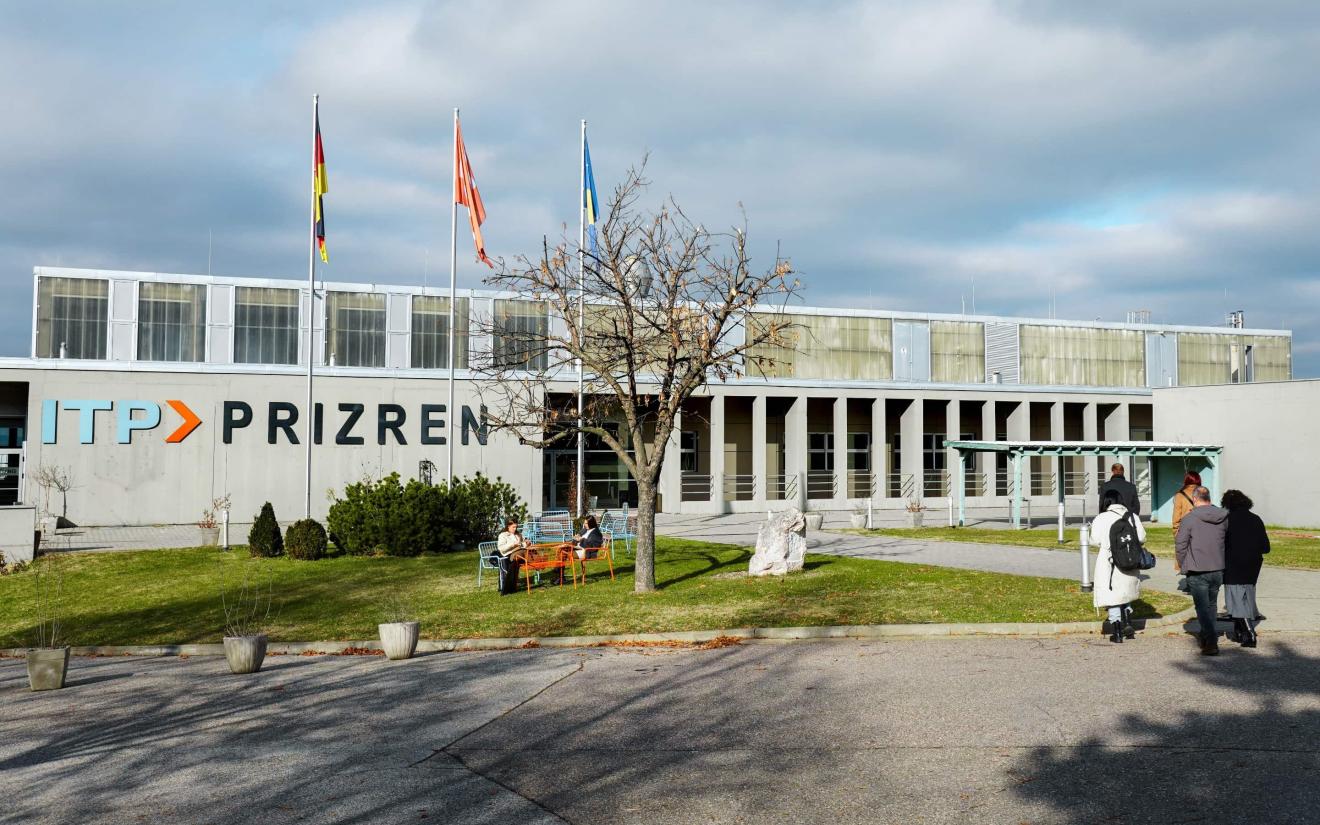 Exterior view of the ITP Prizren building with three flagpoles, seating on the lawn and several people on the grounds.