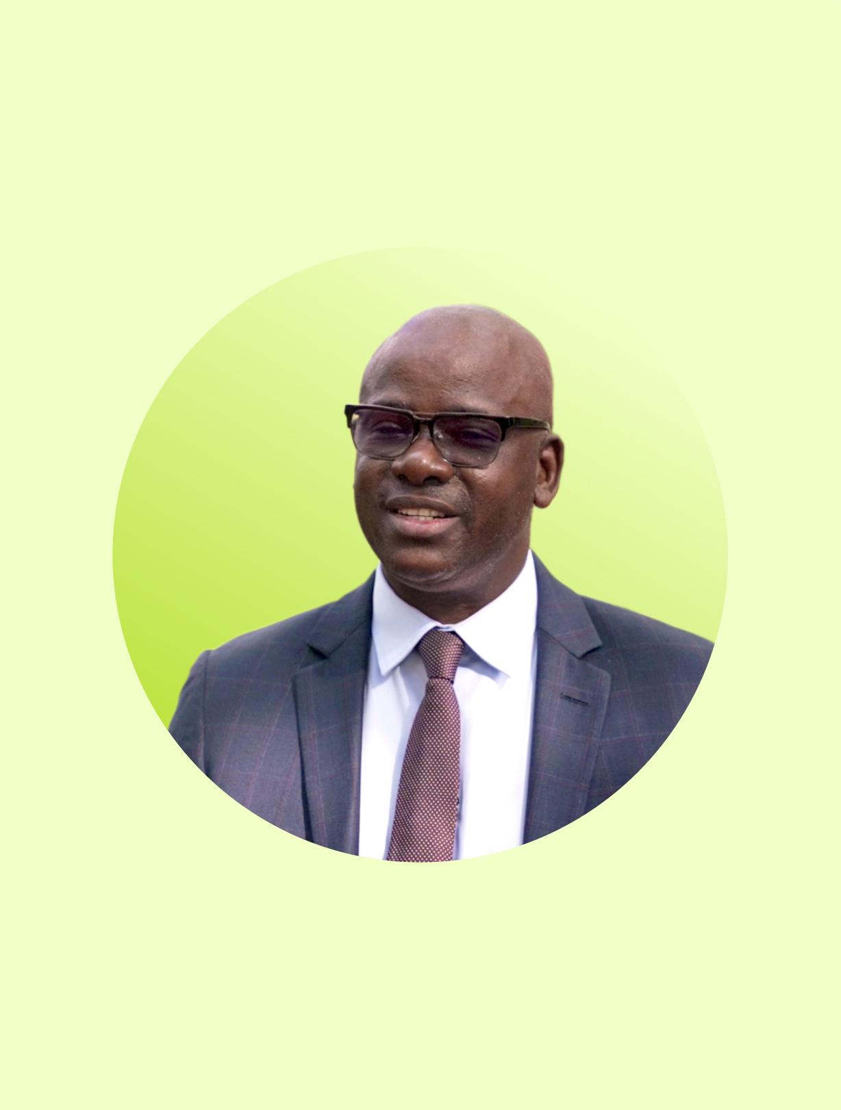 Portrait of a bald man wearing glasses, a dark suit, and a tie against a light green background.