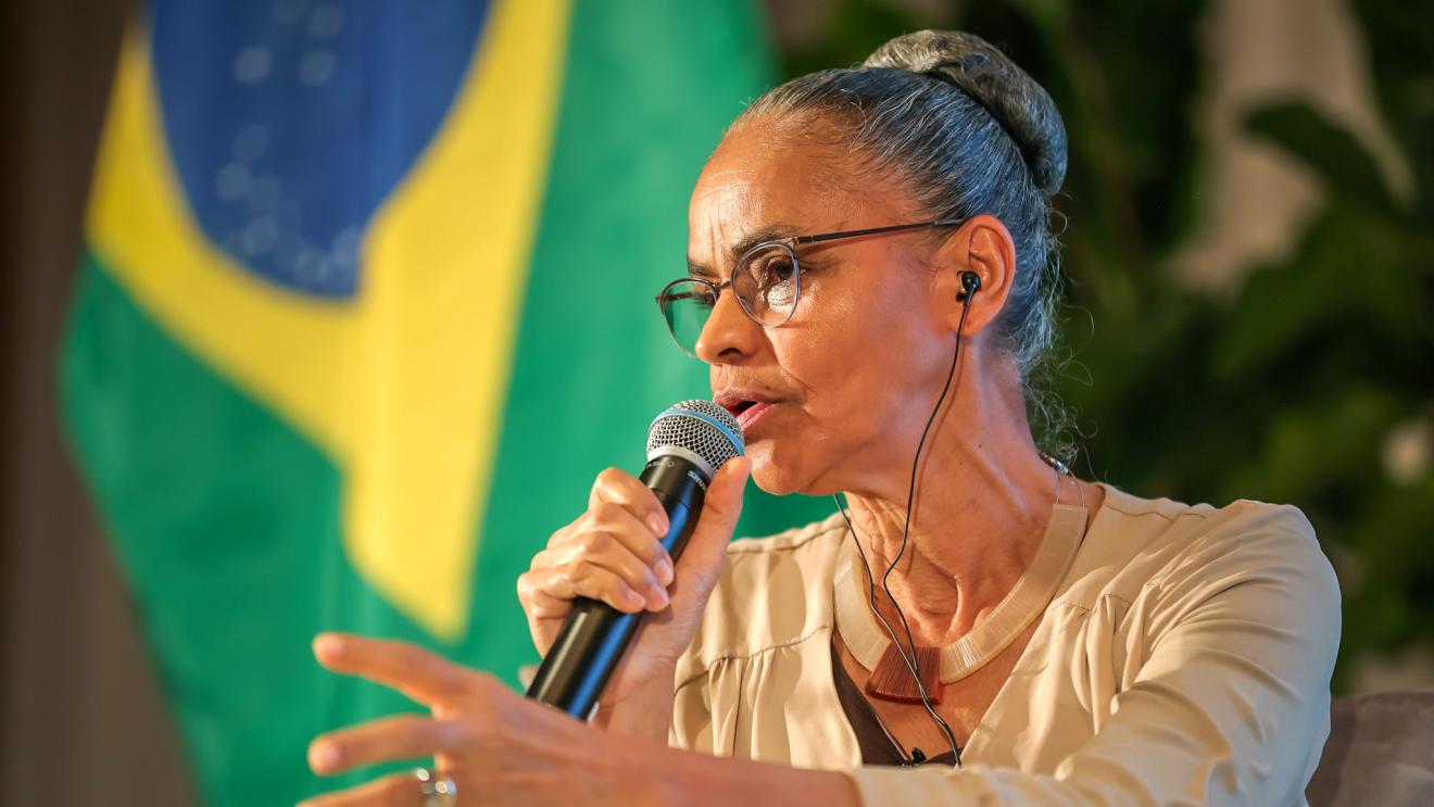 A woman with glasses speaks into a microphone, with the Brazilian flag in the background.
