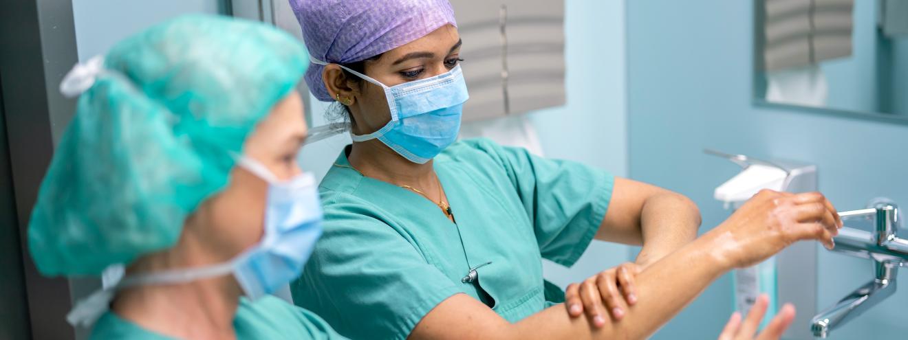Two surgical staff members in green scrubs and face masks carefully disinfect their forearms.