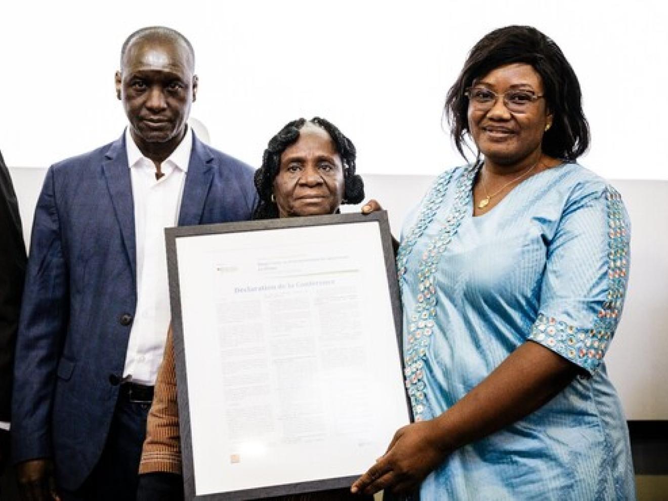 Drei Personen vor einer Wand, eine davon hält einen Bilderrahmen in der Hand