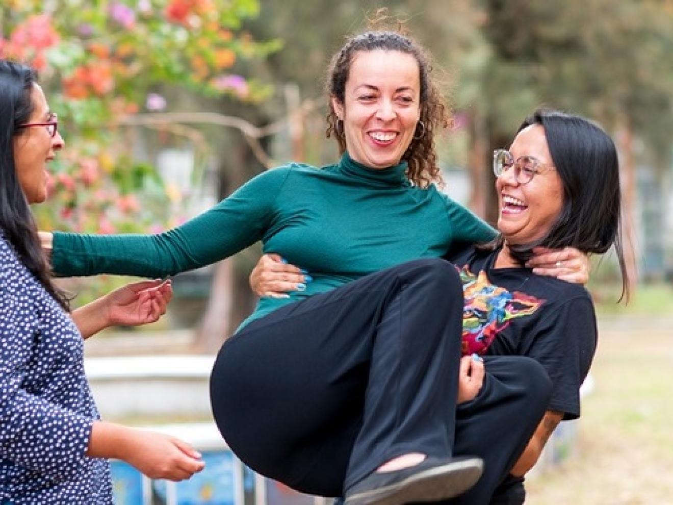 Drei Frauen stehen in einem Kreis beisammen in einem Park. Die Frau rechts hat eine andere Frau auf dem Arm.