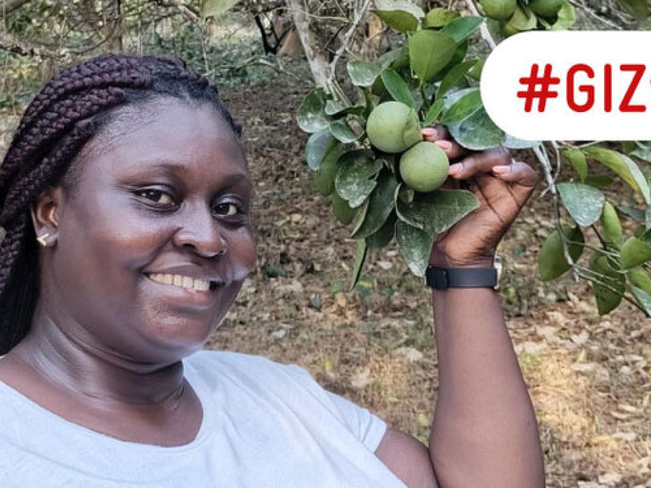 Eine Frau in einem weißen T-Shirt steht lächelnd vor einem Baum. Sie hält einen Zweig mit Limettenfrüchten in der linken Hand. In roter Schrift steht “GIZ wirkt” geschrieben.