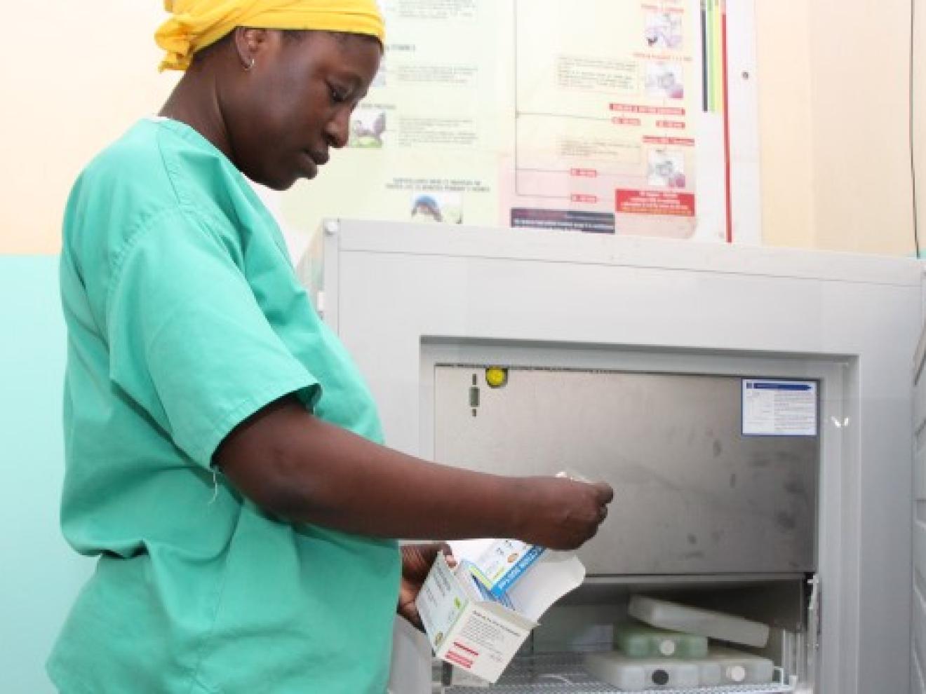 A woman stands in front of an open refrigerator and fills it with medications. She is wearing medical workwear.