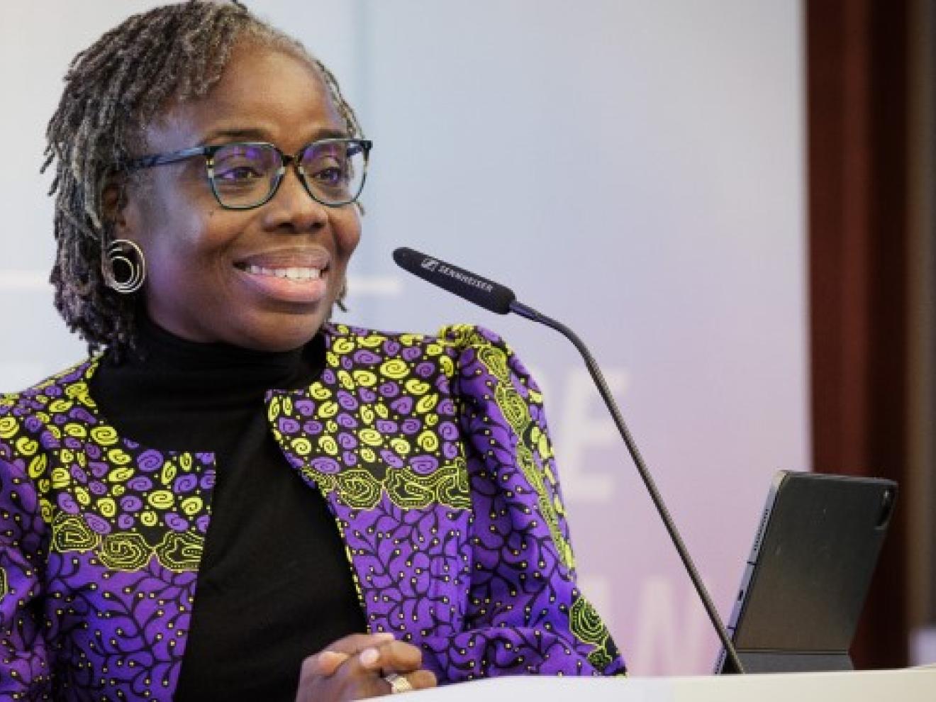 A woman (Mavis Owusu-Gyamfi) in a purple blazer stands at a podium with a microphone and smiles.