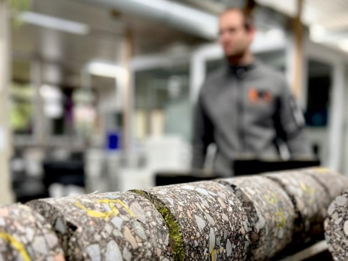 Stücke eines nachhaltigen Straßenbelags im Querschnitt, im Hintergrund eine Fabrik von innen.