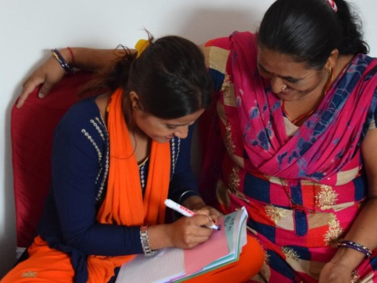 Two women in colorful clothes bend over a notepad together and write something on it.
