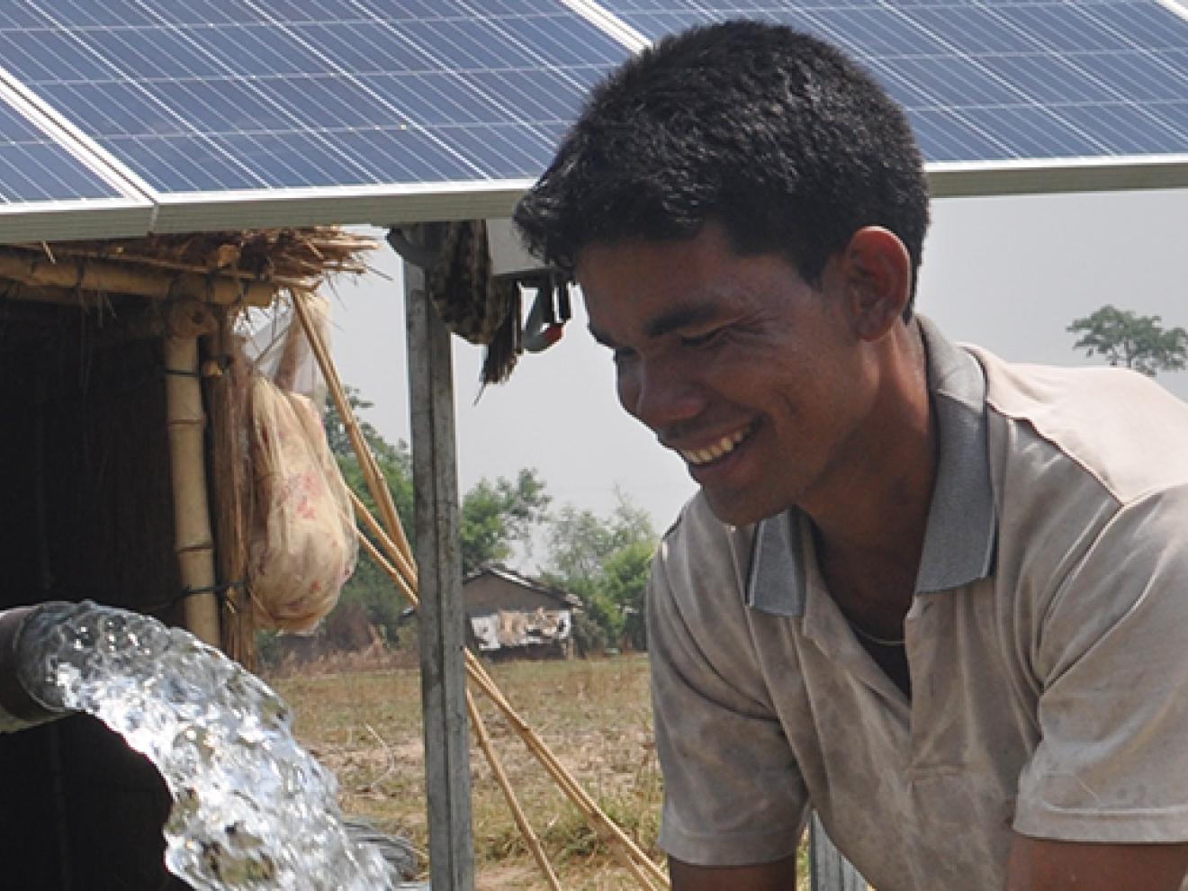 Ein Wasserschwall aus einem Rohr und ein lächelnder Mann vor einer Hütte mit Solarzellen auf dem Dach.