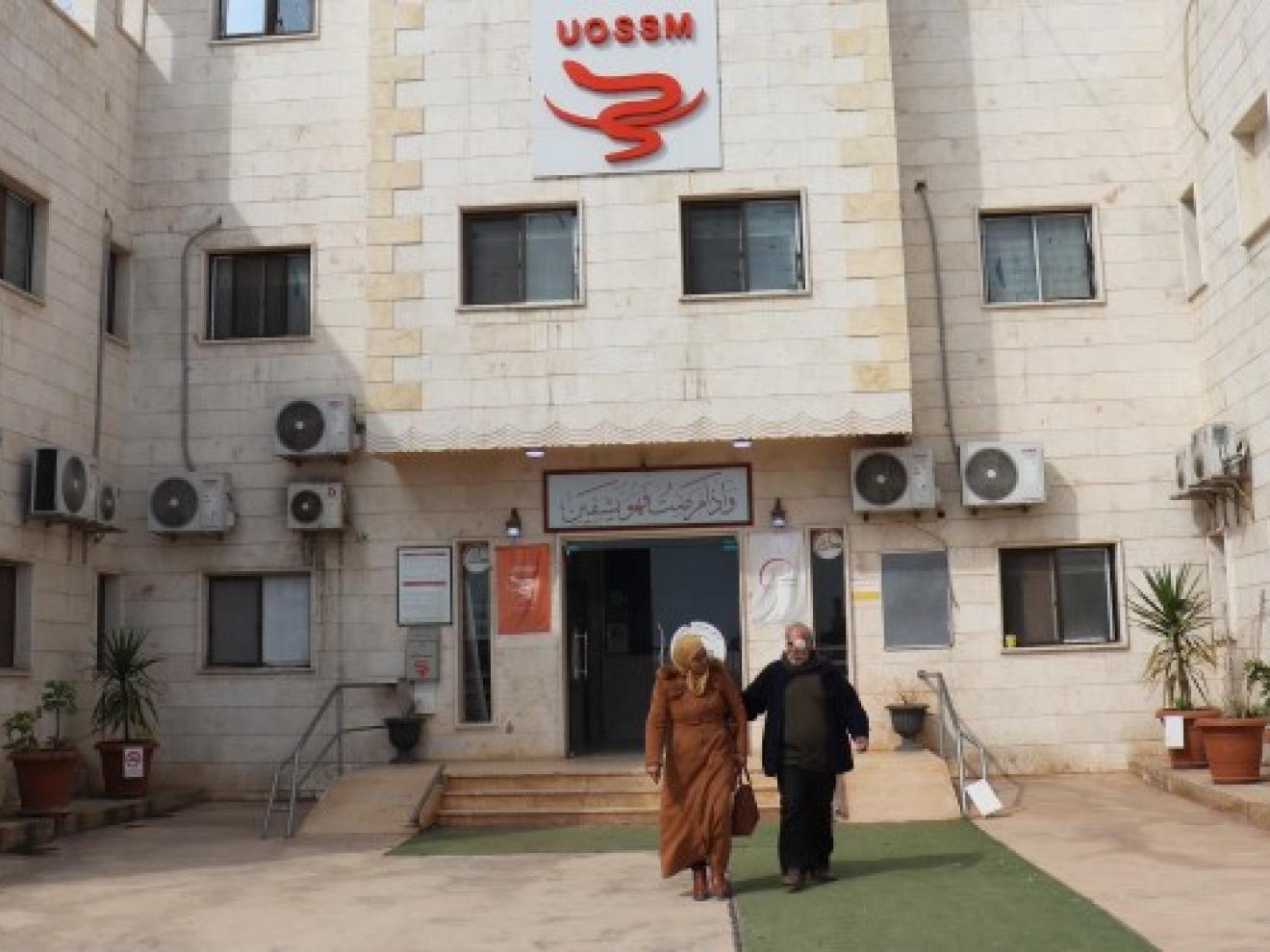 Two people walk in front of a hospital.
