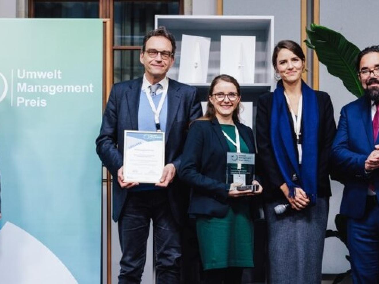 Eine Gruppe von Menschen in Geschäftskleidung steht vor einem grün-weißen Banner mit der Aufschrift ‘Umwelt Management Preis’ und hält eine Glas-Trophäe mit grünem Sockel.