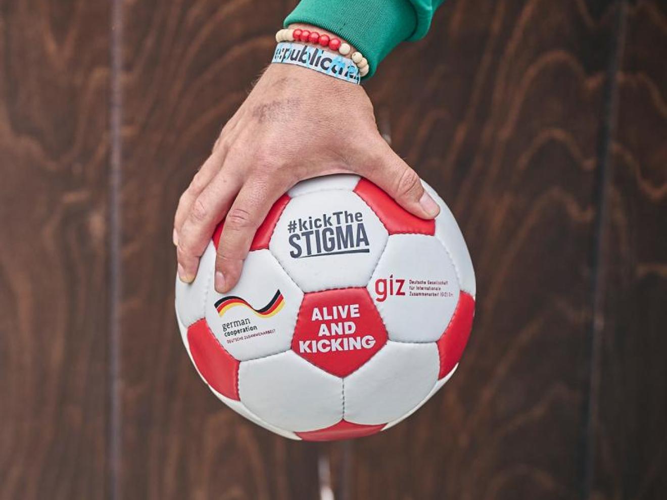 Eine Hand, die einen Fußball ins Bild hält. Aufschrift: #kickTheSTIGMA
