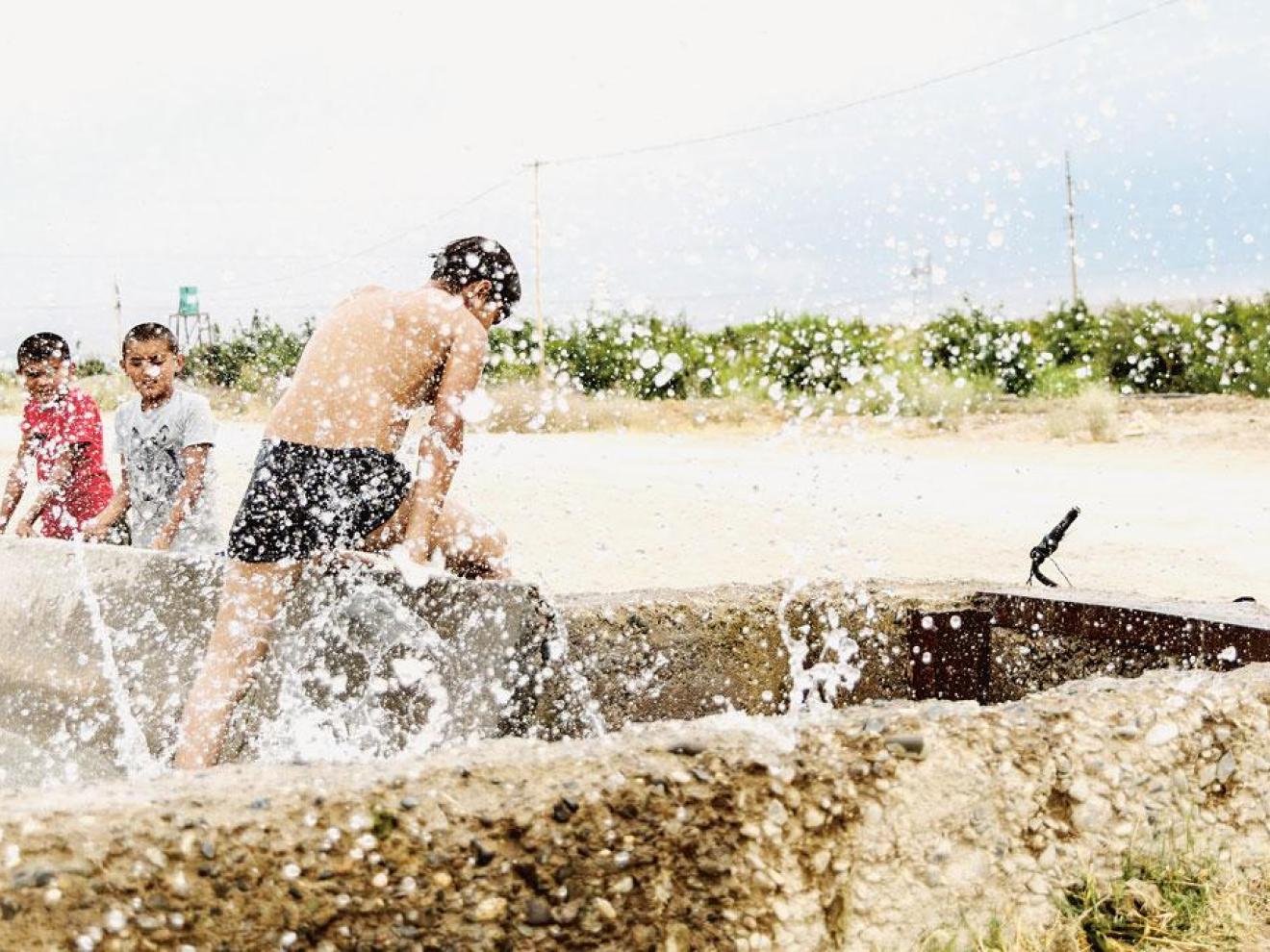 Three children are playing in the water on a sunny day. The child in the foreground is wearing swimwear and actively splashing water, while the other two children, one in pink and the other in grey, are watching. They are located at a water canal in Central Asia.