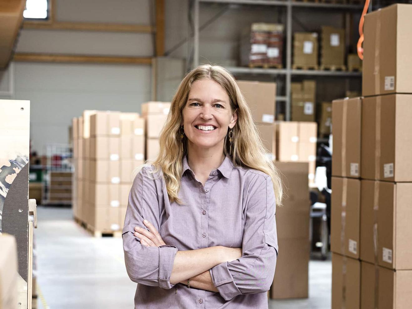 A woman with long blonde hair stands smiling with her arms crossed in a warehouse full of boxes.