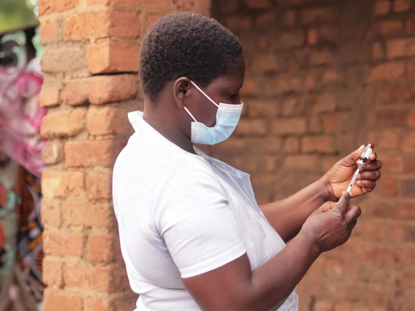 A woman wearing a medical face mask and a white T-shirt draws up an injection from a vaccine vial outdoors.