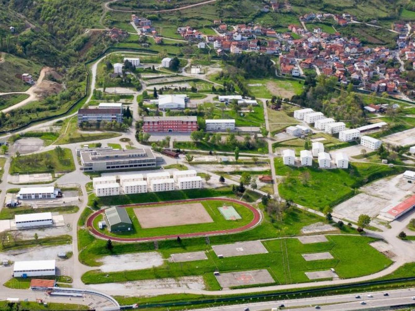 Luftaufnahme eines weitläufigen Geländes mit Gebäuden, Sportanlagen und einer angrenzenden Stadt in einer hügeligen Landschaft.