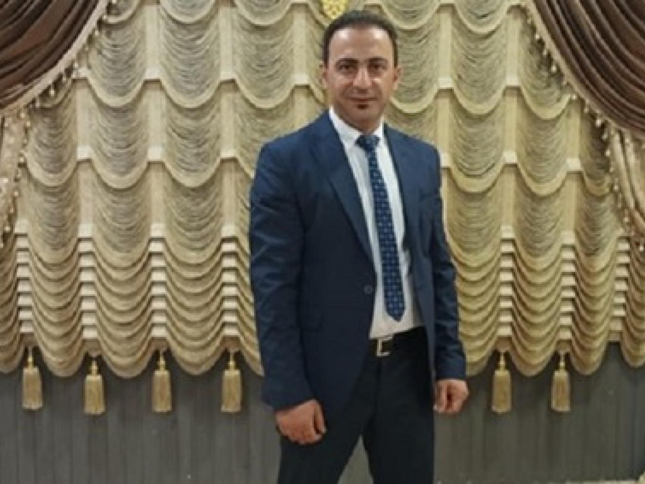 A man wearing a suit and tie is standing in front of artistically draped curtains in gold, beige and brown hues.