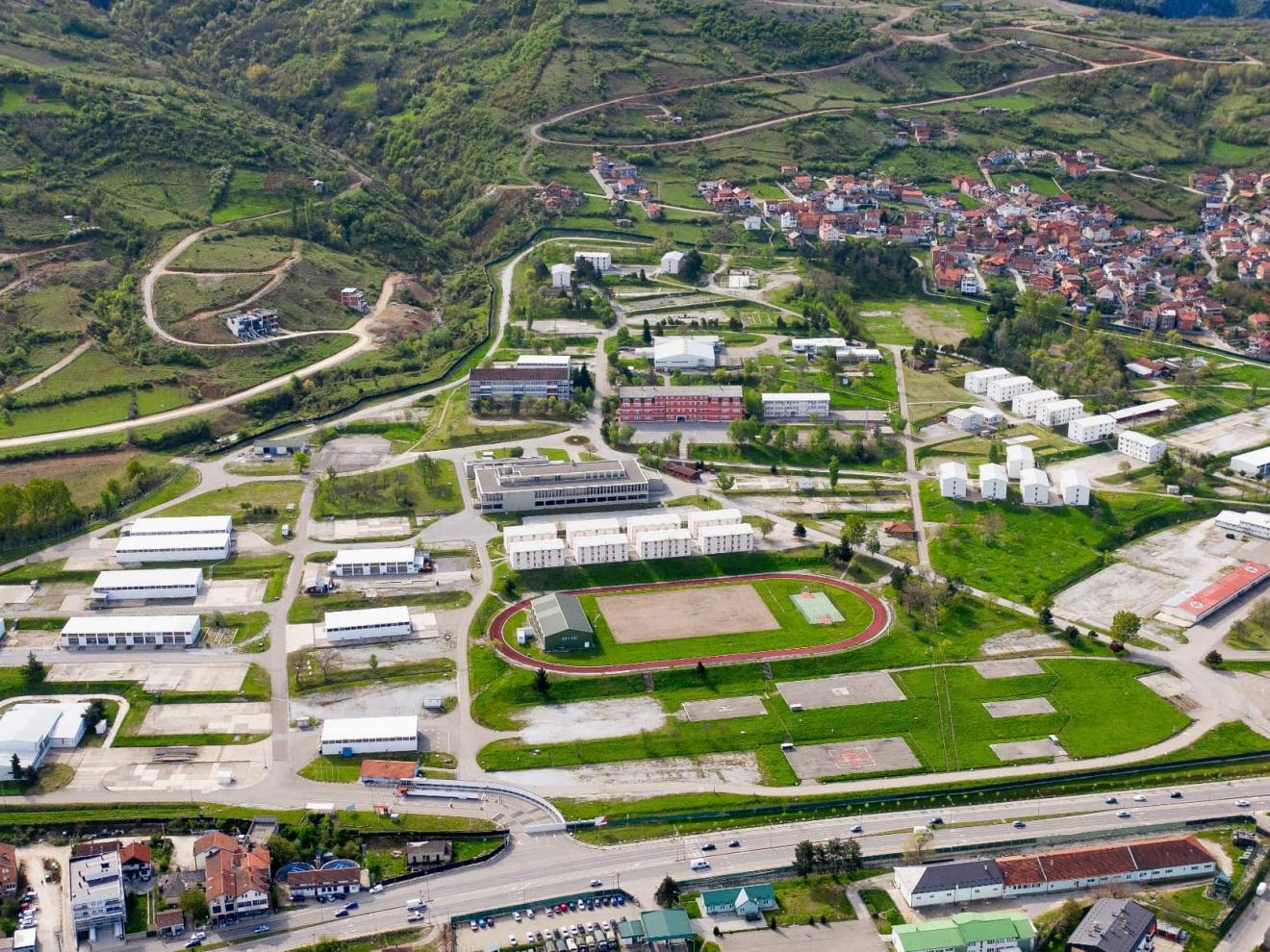 Luftaufnahme eines weitläufigen Geländes mit Gebäuden, Sportanlagen und einer angrenzenden Stadt in einer hügeligen Landschaft.