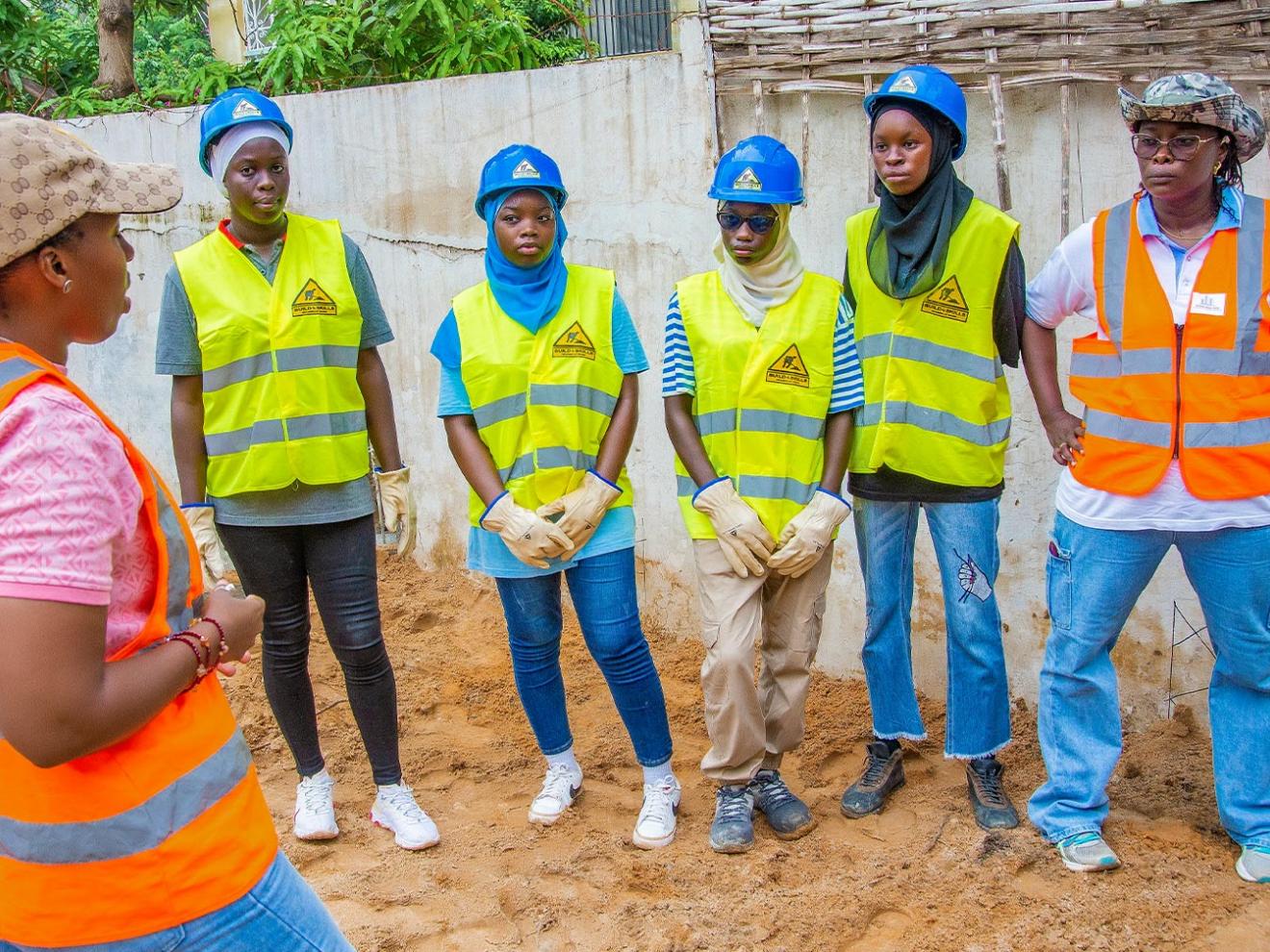 Group photo with Djélia Diagne (left, in profile) talking to five young women wearing protective helmets and high-visibility vests on a construction site.