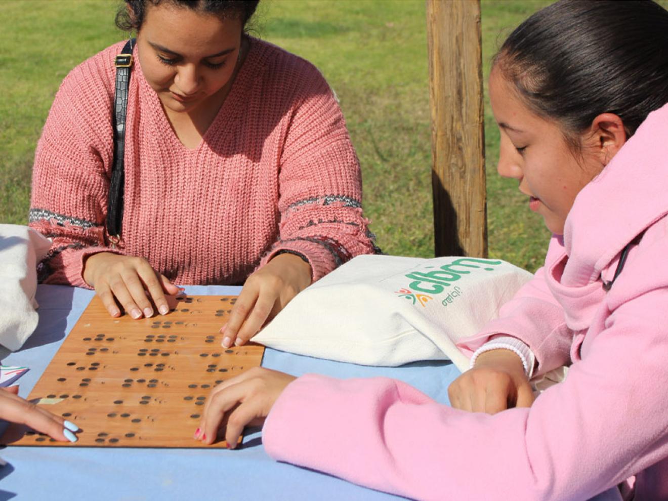 Three young women sitting at a table outdoors run their fingers over a wooden board with Braille.