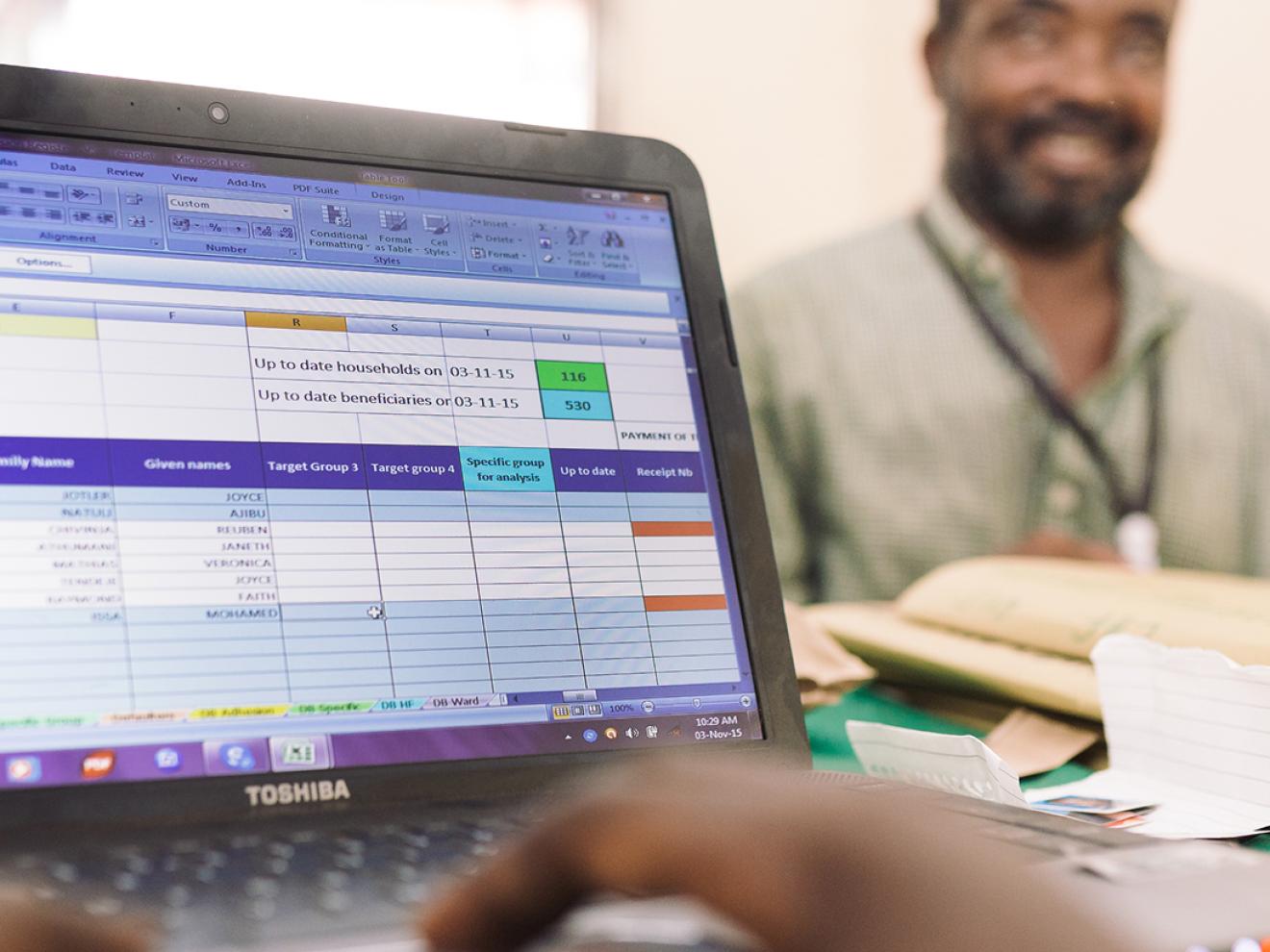 Ein Mensch tippt auf einem Laptop mit Excel-Tabelle, im Hintergrund sitzt ein lächelnder Mann an einem Schreibtisch.