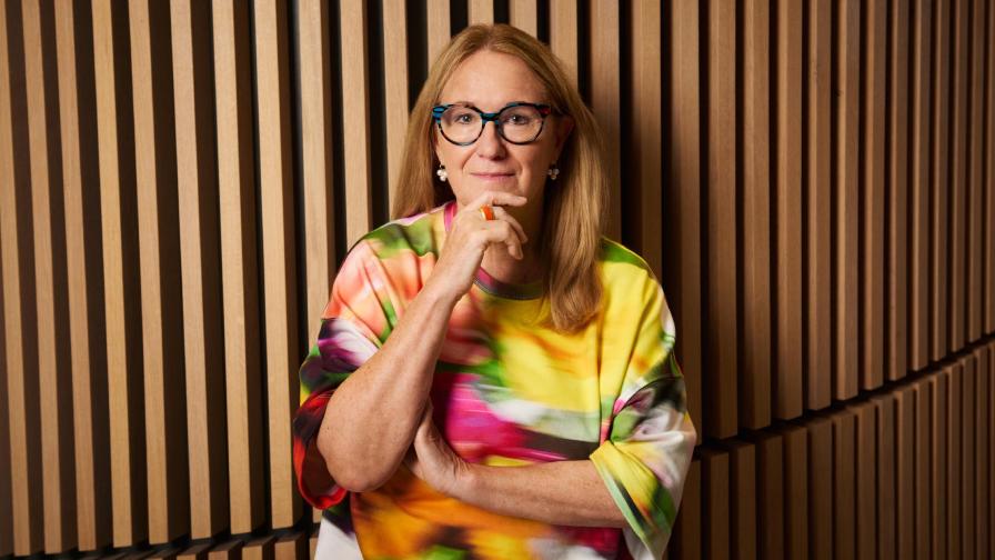 A woman with long blonde hair and glasses stands in front of a wall with wooden slats and wears a colourful blouse.