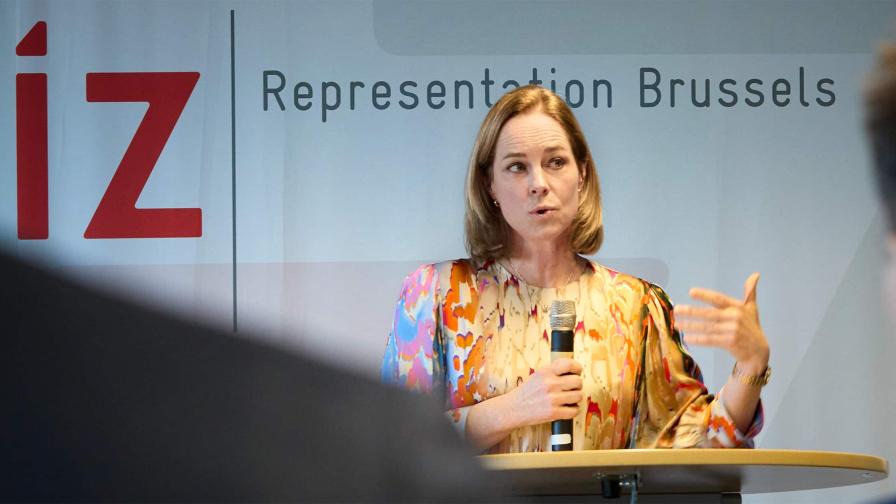  Eine Frau mit schulterlangem blondem Haar trägt eine farbenfrohe Bluse mit abstraktem Muster und spricht auf einer Veranstaltung in Brüssel an einem Rednerpult mit einem Mikrofon in der Hand.