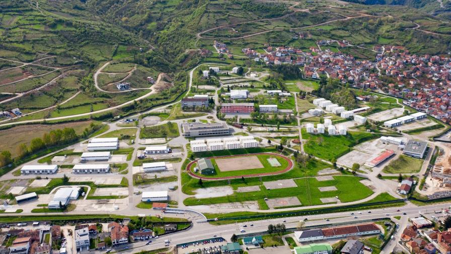 Luftaufnahme eines weitläufigen Geländes mit Gebäuden, Sportanlagen und einer angrenzenden Stadt in einer hügeligen Landschaft.
