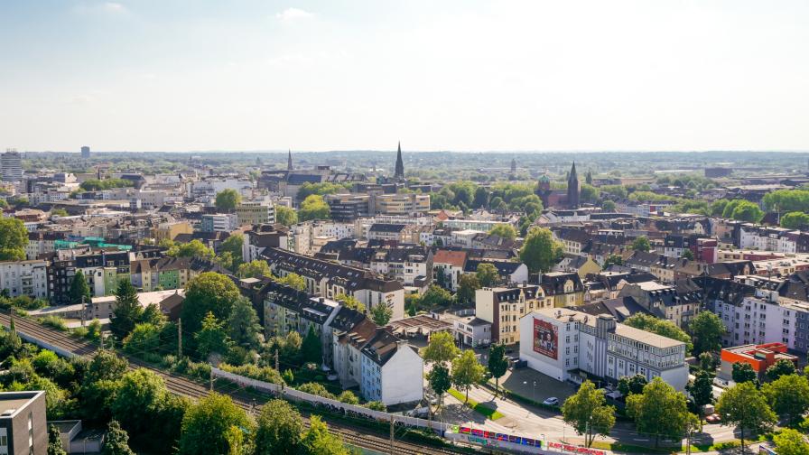 Luftaufnahme einer Stadt mit dicht bebauten Wohnhäusern, mehreren Kirchtürmen und Bahngeleisen im Vordergrund, umgeben von vielen Bäumen und Grünflächen.