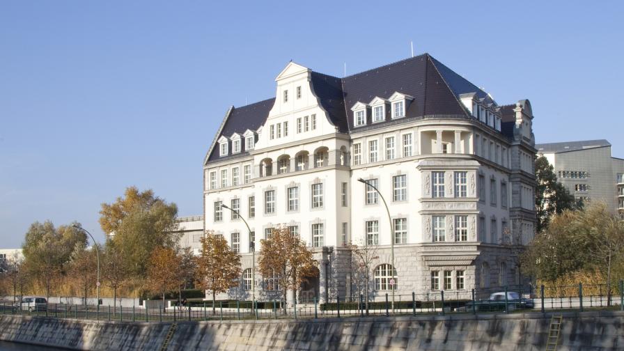 Historisches, mehrstöckiges Gebäude mit dunklem Schrägdach und zahlreichen Fenstern am Ufer eines Kanals; umgeben von Bäumen und modernen Gebäuden im Hintergrund, bei klarem Himmel.