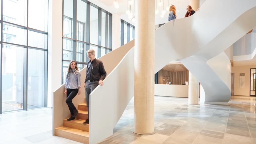 Heller und hoher Innenraum eines modernen Gebäudes mit einer Säule und einem organischen Treppenaufgang. Auf der Treppe laufen vier Personen.