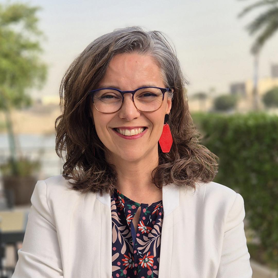 Porträt einer lächelnden Frau mit schulterlangem, braun-grauem Haar, rotem Ohrring, Brille und weißem Blazer vor einem unscharfen, grünen Außenhintergrund.