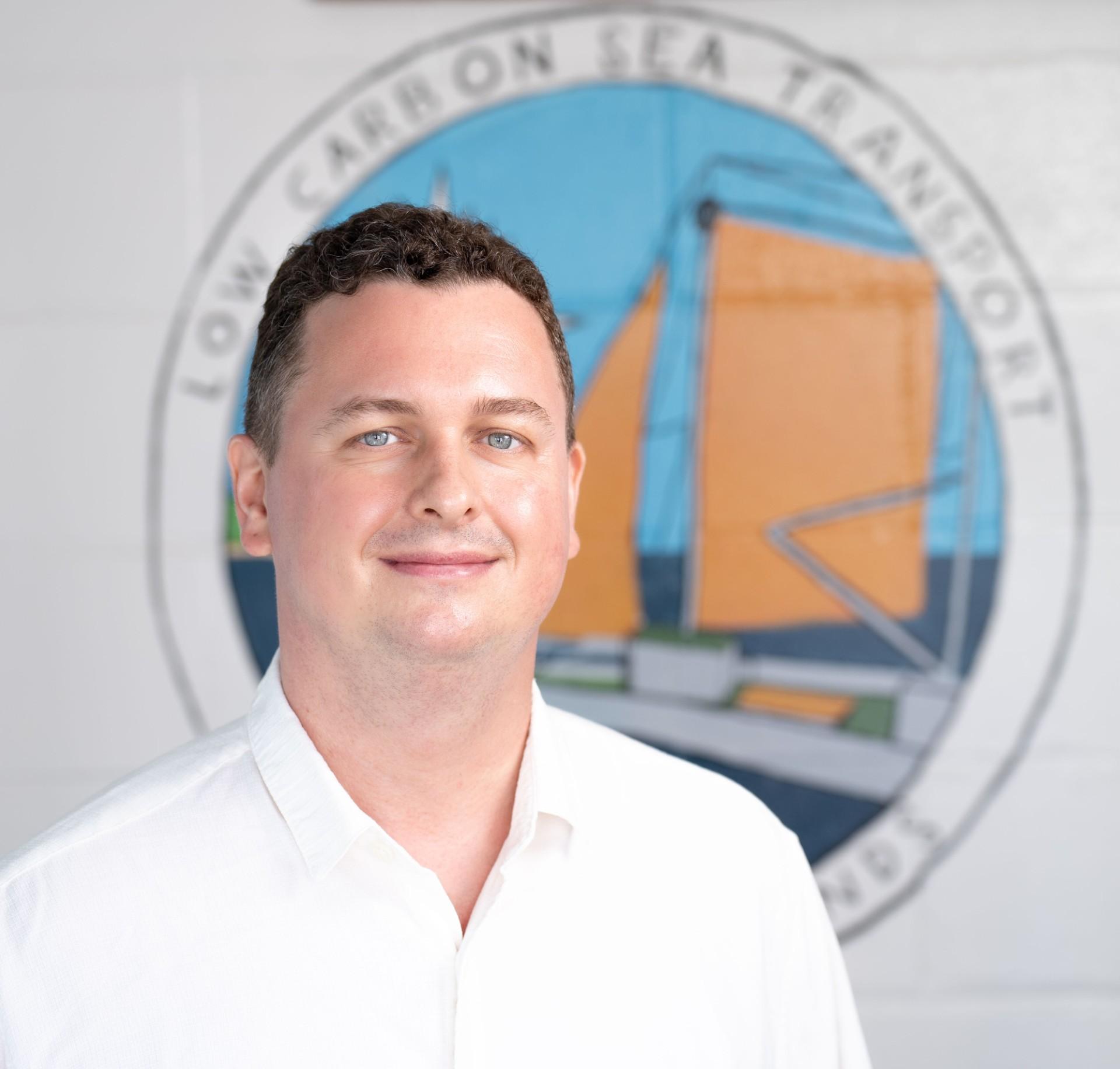 Porträt eines lächelnden Mannes mit kurzen braunen Haaren in weißem Hemd vor einem Wandbild mit der Aufschrift „Low Carbon Sea Transport“ und einer stilisierten Segelbootgrafik.