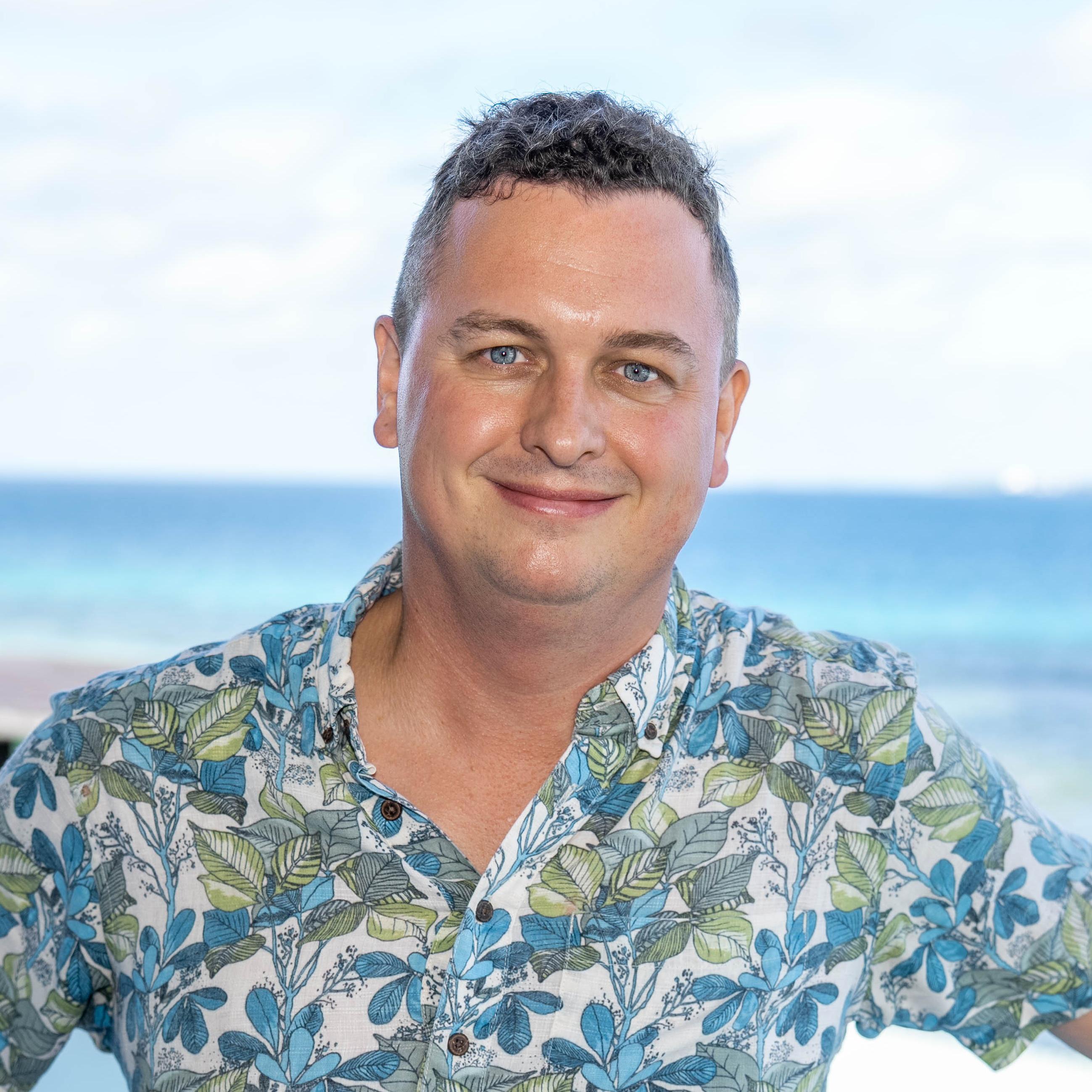 Ein lächelnder Mann mit kurz gelocktem Haar trägt ein buntes Hemd mit Blattmuster und steht vor einem unscharfen Hintergrund mit Meer und Himmel.