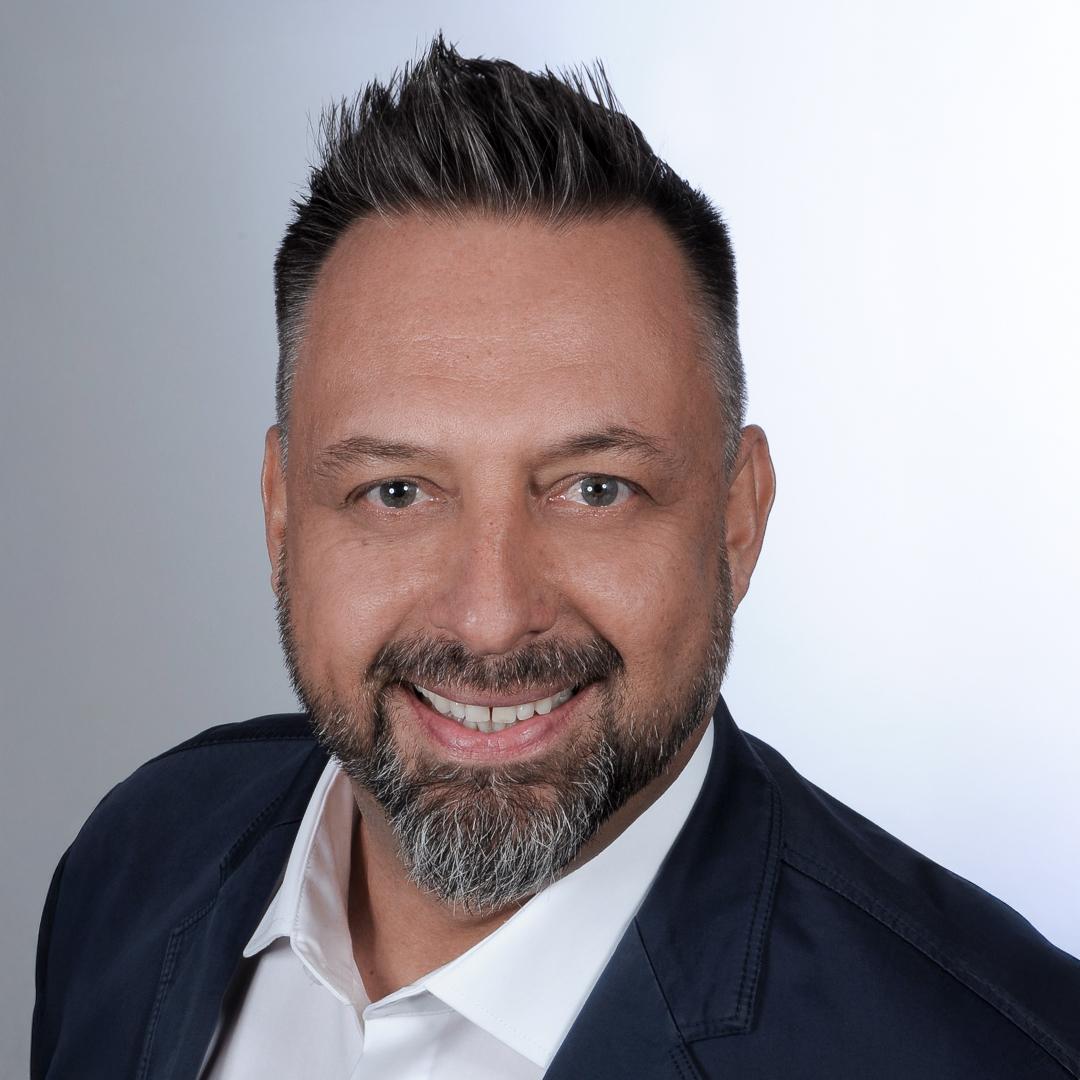 Porträt eines Mannes mit kurzem, grauem Haar und einem gepflegten Bart. Er trägt ein weißes Hemd und eine dunkle Jacke. Der Mann lächelt freundlich in die Kamera, vor einem neutralen, hellen Hintergrund.