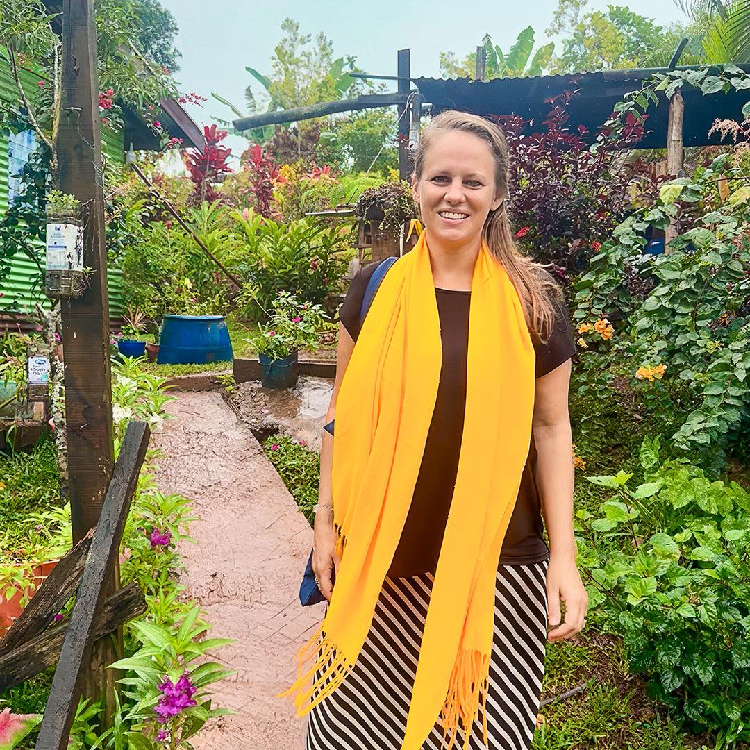 Eine lächelnde Frau mit gelbem Schal steht auf einem schmalen Weg in einem üppig grünen Garten mit vielen Pflanzen und Blumen.