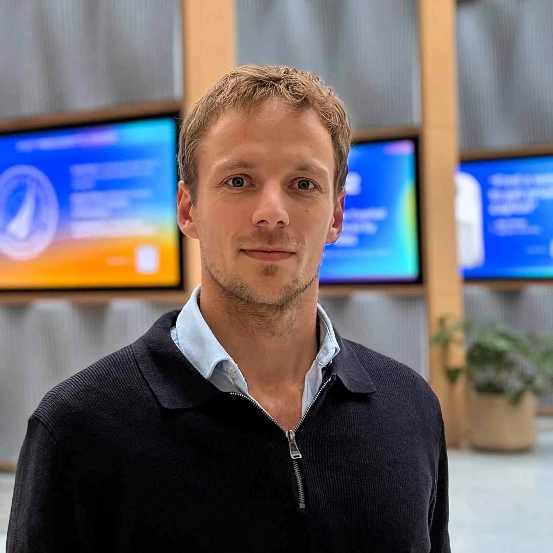 Jonas Wittern - Porträtfoto eines Mannes in einem Innenraum, der in die Kamera blickt, mit unscharfen Bildschirmen im Hintergrund.