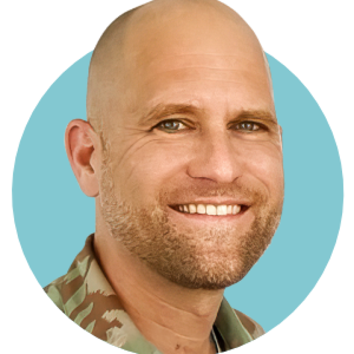 Ein Porträt eines lächelnden, kahlköpfigen Mannes in militärischer Tarnuniform vor einem kreisförmigen blauen Hintergrund.