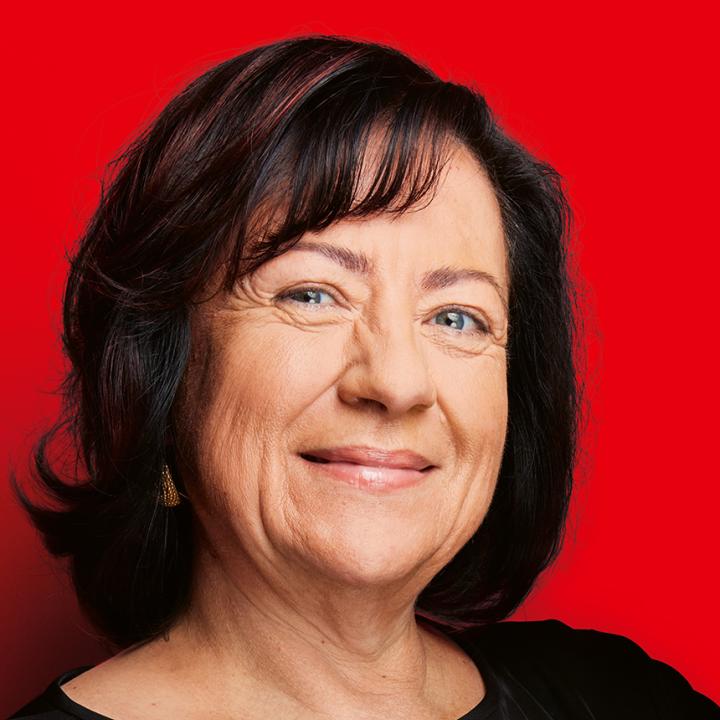 Porträt einer lächelnden älteren Frau mit dunklem, schulterlangem Haar und schwarzem Oberteil vor knallrotem Hintergrund.