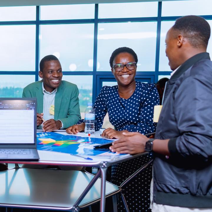 Several people sit around a table in a bright room with a laptop and documents, talking, with a “One Health Data Alliance Africa” banner in the background.