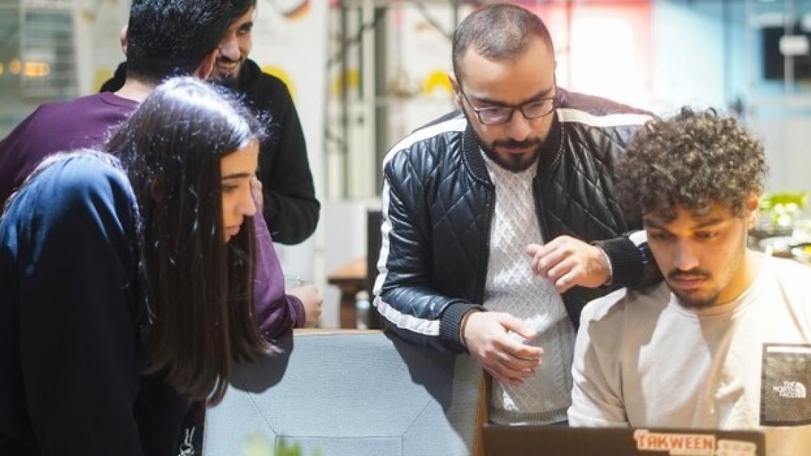 Five young people in a modern room; three look at a laptop while two chat.