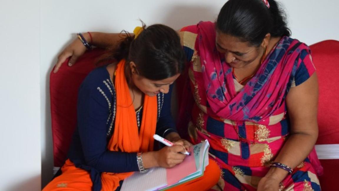 Zwei Frauen in bunter Kleidung beugen sich gemeinsam über einen Notizblock und schreiben etwas darauf.