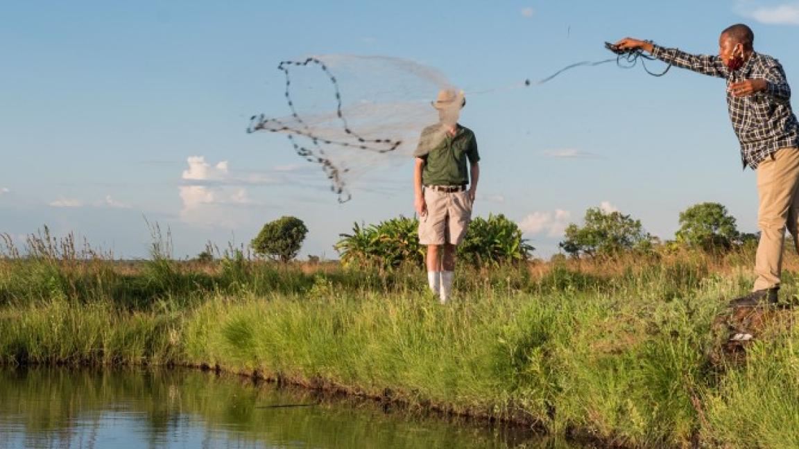 A man at a pond throws out a fishing net; a second man is in the background.