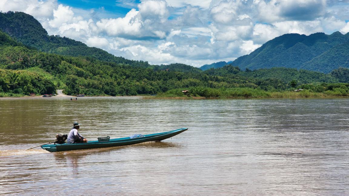 A man is sailing his boat on the Mekong River. In the background are forested mountains.