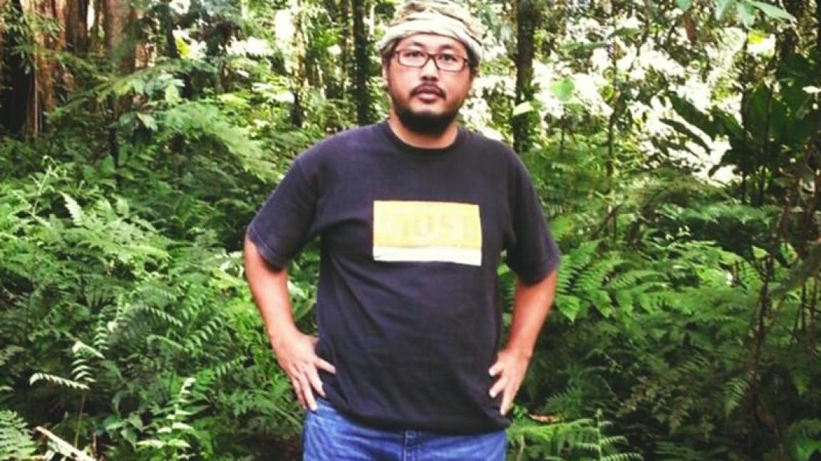 Ein Mann mit Brille, schwarzem T-Shirt und Jeans steht inmitten eines dichten, tropischen Regenwaldes mit üppigem Farnbewuchs und hohen Bäumen.