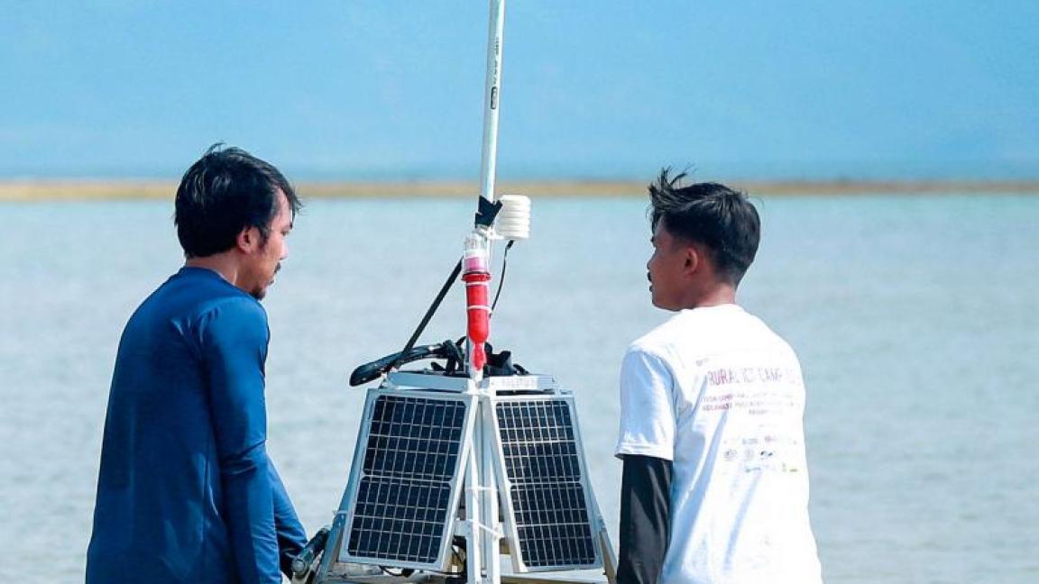 Zwei Männer tragen eine solarbetriebene Messboje durch flaches Wasser, im Hintergrund sind Berge und eine Sandbank zu sehen.