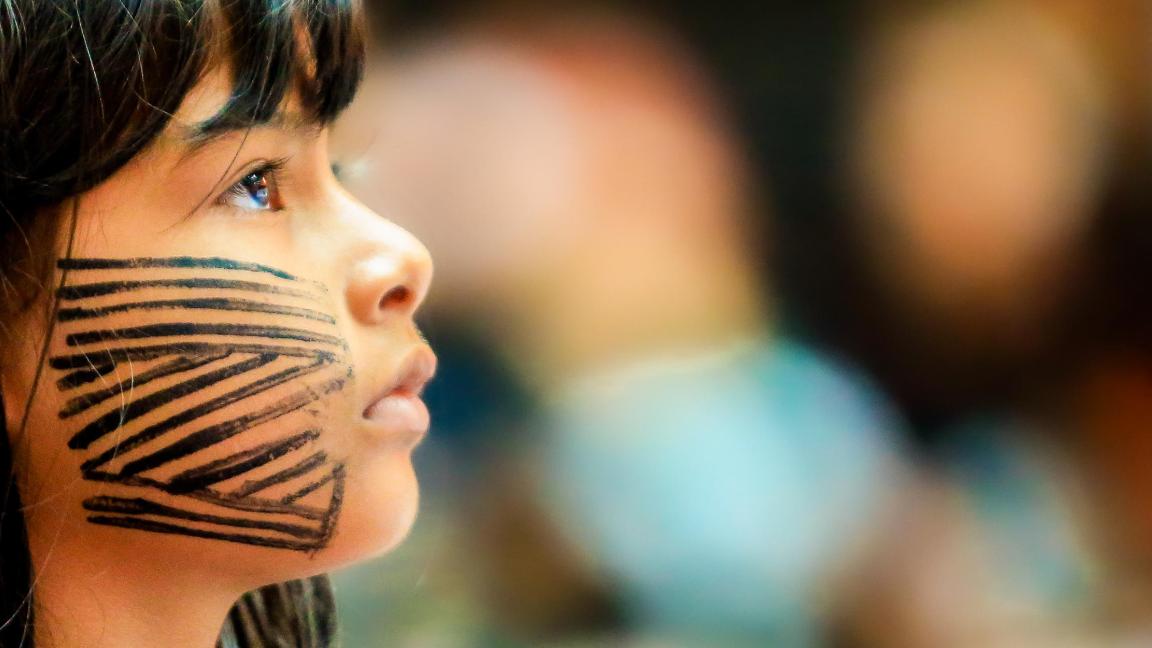 Nahaufnahme eines Kindes mit schwarzen Streifenmustern im Gesicht, das zur Seite blickt, mit verschwommenem Hintergrund.