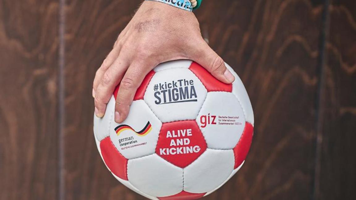 Eine Hand, die einen Fußball ins Bild hält. Aufschrift: #kickTheSTIGMA
