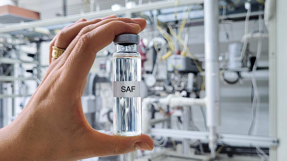 Eine Hand hält ein kleines, mit "SAF" beschriftetes Fläschchen mit klarer Flüssigkeit vor einem verschwommenen industriellen Hintergrund.