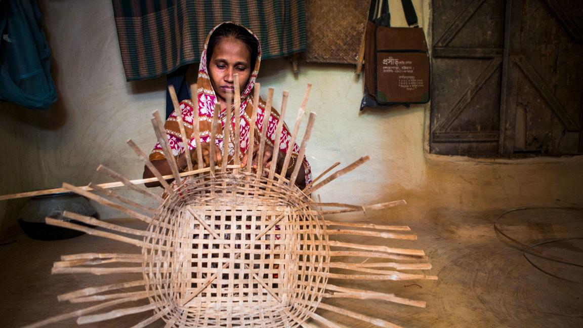 Eine Frau in traditioneller Kleidung sitzt in einem einfachen Innenraum und flechtet konzentriert einen grossen Korb aus Bambus oder ähnlichem Naturmaterial.       ChatGPT fragen